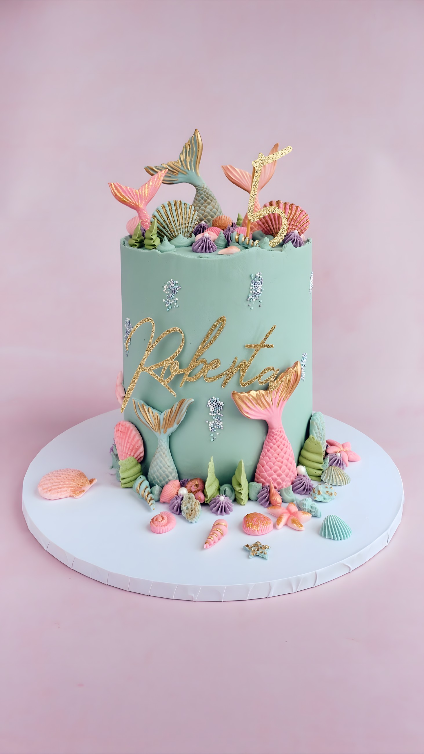 A tall mint-green buttercream cake decorated with pastel mermaid tails, seashells and a gold name topper
