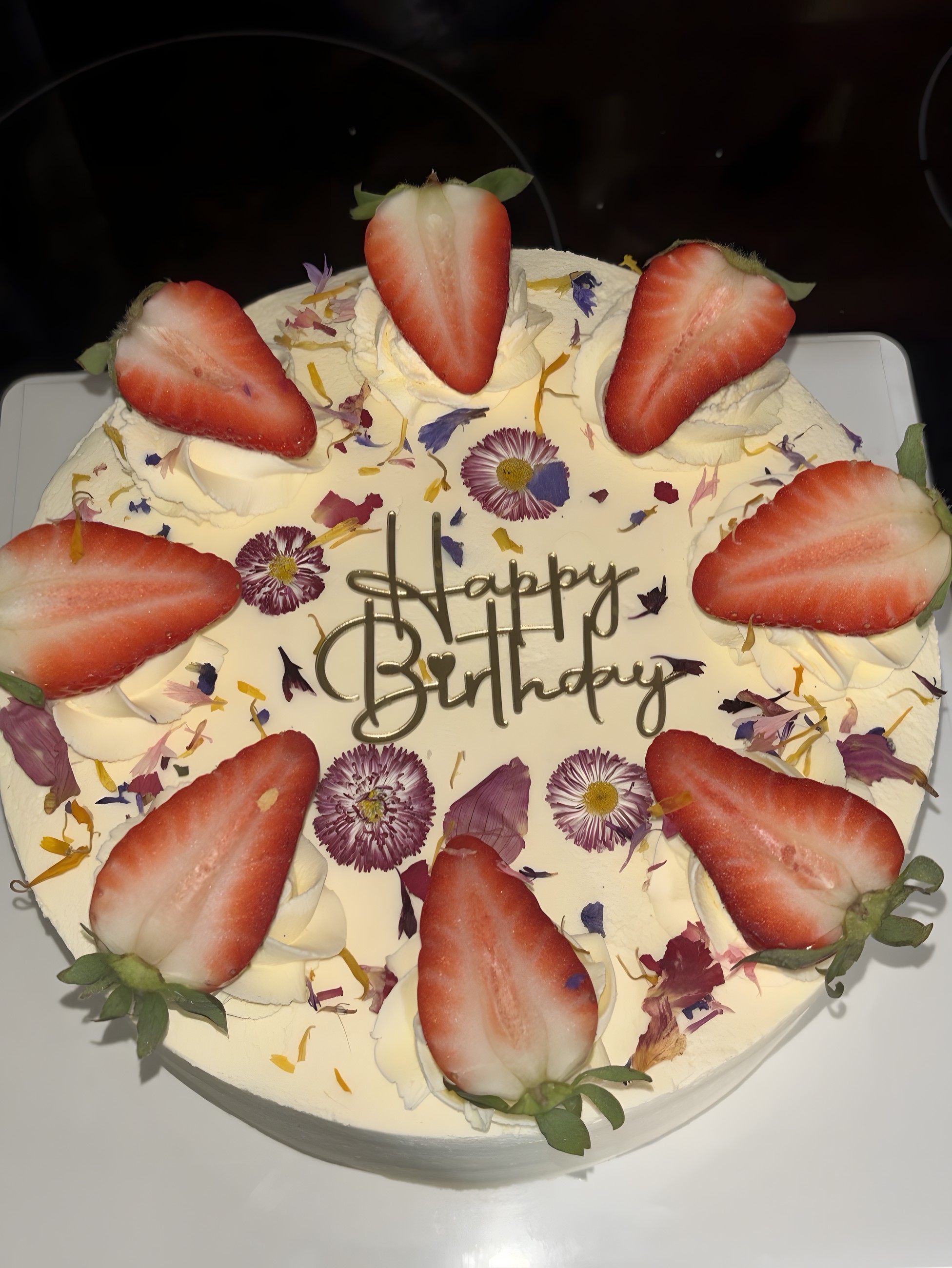 A white cream birthday cake ringed with fresh strawberry halves and pressed edible flowers